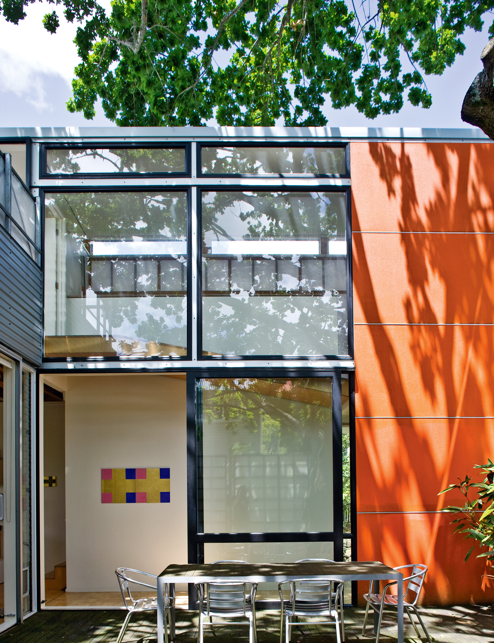 The outdoor dining area stands before the double-height entry foyer (with its timber ramp leading to the upper floor), and its striking metal framing and orange wall.